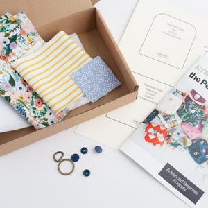 May include: An open cardboard box filled with fabric pieces in floral, striped, and dotted patterns. The box also contains a sewing pattern, a booklet titled "the Petite Case", and various hardware components like buttons and a key ring.