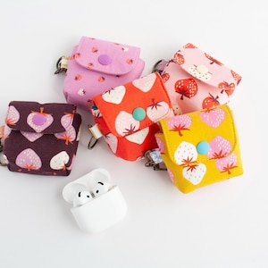 May include: A collection of colorful fabric cases for wireless earbuds, featuring various strawberry patterns in pink, red, and yellow. Each case has a snap closure and a small metal clasp. A white earbud charging case is also present.