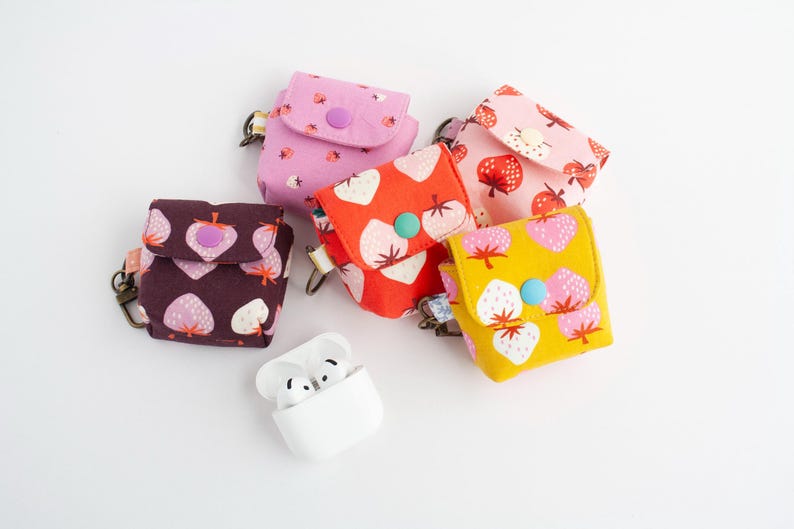May include: A collection of colorful fabric cases for wireless earbuds, featuring various strawberry patterns in pink, red, and yellow. Each case has a snap closure and a small metal clasp. A white earbud charging case is also present.