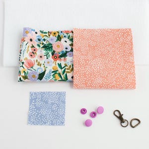 May include: A collection of fabric squares and hardware for crafting. Includes a floral print, a peach dotted print, and a blue dotted print. Also shown are pink snaps and bronze-colored metal clasps.