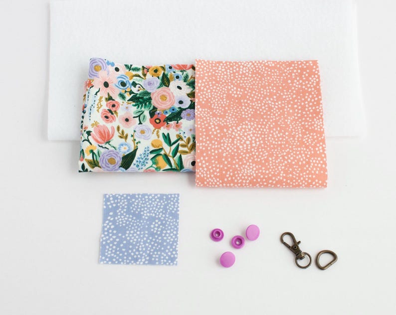May include: A collection of fabric squares and hardware for crafting. Includes a floral print, a peach dotted print, and a blue dotted print. Also shown are pink snaps and bronze-colored metal clasps.