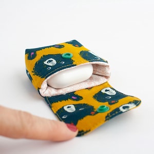 May include: A yellow and teal fabric case with a bear pattern, holding a white earbud. The case is open, revealing a pink patterned interior and green snap closures. The case is designed to protect earbuds.