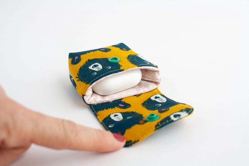 May include: A yellow and teal fabric case with a bear pattern, holding a white earbud. The case is open, revealing a pink patterned interior and green snap closures. The case is designed to protect earbuds.