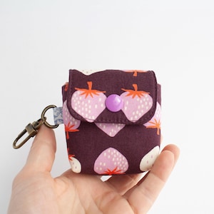 May include: A small, square fabric pouch with a flap closure, featuring a repeating pattern of pink strawberries with orange stems on a dark burgundy background. It has a purple button closure and a metal clasp.