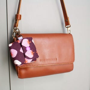 May include: A brown leather crossbody bag with a matching strap and a small, patterned pouch attached. The pouch features a dark purple background with a strawberry print and a pink button closure. The bag is embossed with "PORTLAND LEATHER".