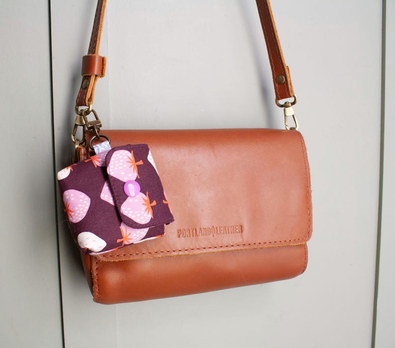 May include: A brown leather crossbody bag with a matching strap and a small, patterned pouch attached. The pouch features a dark purple background with a strawberry print and a pink button closure. The bag is embossed with "PORTLAND LEATHER".