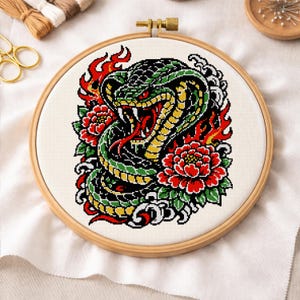 May include: Cross-stitch embroidery in a wooden hoop. The design features a green, black, and yellow snake with red eyes and tongue, surrounded by red flowers and flames. The background is off-white.