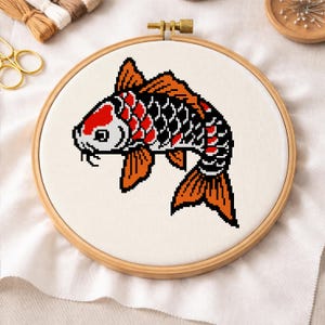 May include: A cross-stitch embroidery of a koi fish in a wooden hoop. The fish features orange, red, black, and white colors. The embroidery is on a cream-colored fabric. The hoop is secured with a brass clasp. The theme is Japanese art.