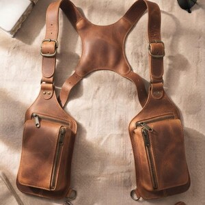 May include: A brown leather chest harness featuring two symmetrical pouches with zippered closures. The harness includes adjustable shoulder straps with metal buckles and a central back panel, designed to be worn over the torso for storage.
