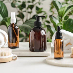 May include: An assortment of amber glass bottles with black pumps and sprayers, and a dropper bottle, displayed on a white surface. A wooden handled brush, white towels, and a woven basket are also present, suggesting a spa or self-care theme.