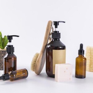 Könnte beinhalten: Eine Auswahl an braunen Glasflaschen mit schwarzen Pumpen und Pipetten, eine Holzbürste, ein Seifenstück und ein Naturschwamm. Eine kleine Topfpflanze ist ebenfalls vorhanden, was auf natürliche Hautpflegeprodukte hindeutet.