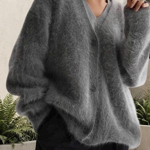Può includere: Un cardigan grigio antracite con scollo a V, maniche lunghe e chiusura frontale con bottoni. Il capo presenta una finitura in maglia spazzolata morbida e testurizzata, indossato sopra una maglia grigio chiaro con scollo a V e pantaloni scuri.