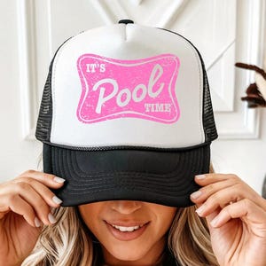May include: A trucker hat with a white front panel and black mesh back. The front features a pink, distressed-style rectangular graphic with rounded corners containing the white text 'IT'S Pool TIME'. The hat has a solid black brim and a black cord.