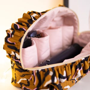 May include: Open cosmetic bag with a brown, black, and white patterned exterior and a pink interior. The bag contains a hair dryer, brushes, and other beauty accessories. The bag has a ruffled edge.