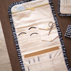 Op de afbeelding: Een crèmekleurige stoffen opberghoes met een zwart-witte gingham ruche-rand. De binnenkant is voorzien van een ritsvak, meerdere verticale sleuven voor breinaalden, twee zakjes met drukknopen en een goudkleurige ritstrekker.