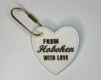 May include: A heart-shaped white fabric patch with a gold-toned metal lobster clasp attached. The patch features dark brown embroidered text that reads FROM Hoboken WITH LOVE, with a white stitched border around the entire heart shape.