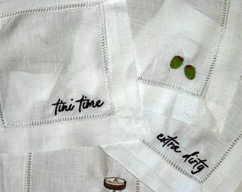 May include: Three white square linen cocktail napkins featuring hemstitched borders. One napkin is embroidered with the black text "tini time", another with two green olives, and the third with a black martini glass and the text "extra dirty".