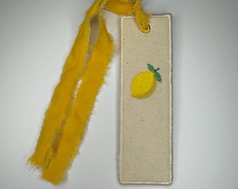 May include: A rectangular off-white fabric bookmark featuring an embroidered yellow lemon with a green leaf. The bookmark has a stitched border and a gold-toned metal eyelet at the top, through which a long, frayed yellow ribbon is tied.