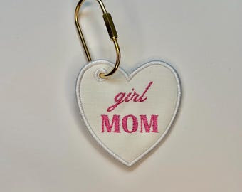 May include: A heart-shaped keychain with the words "girl MOM" embroidered in pink on a white background. The keychain has a gold-colored clasp for attaching to keys or a bag.