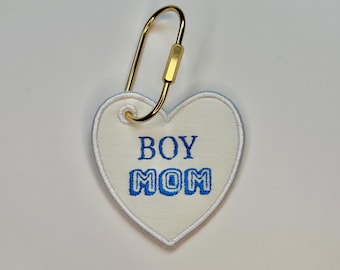 May include: A heart-shaped keychain with the embroidered text "BOY MOM" in blue. The keychain has a gold-colored carabiner clip for attachment.