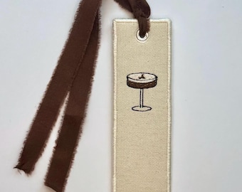 May include: A beige fabric bookmark with a brown embroidered cocktail illustration. The bookmark has a brown ribbon attached at the top. The edges of the bookmark are stitched with a cream-colored thread.