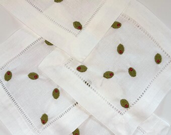 May include: A set of square white linen napkins featuring an embroidered pattern of green olives with small red pimentos. Each napkin has a decorative hemstitched border detail along the edges, creating a uniform look across the collection.