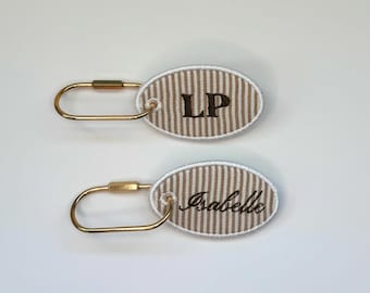 May include: Two oval-shaped fabric tags with a beige and white vertical striped pattern. Each tag features a gold-toned metal wire loop attached through a grommet. One tag is embroidered with the letters LP, and the other with the name Isabelle.