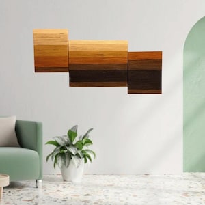 May include: A contemporary wall art piece featuring three wooden blocks in shades of brown. The blocks are arranged horizontally, creating a gradient effect. The artwork is mounted on a white wall. The piece would look great in a living room.