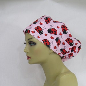 May include: A pink scrub cap featuring a repeating pattern of red ladybugs and red hearts. The ladybugs have black spots and antennae. The cap is made of a soft fabric and is designed to cover the hair.