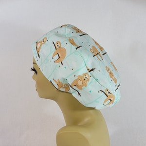 May include: A light blue surgical scrub cap featuring a repeating pattern of brown sloths hanging from tree branches. The cap is made of a soft fabric and is designed to cover the hair during medical procedures or other activities.