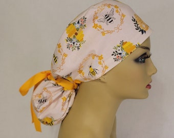 May include: A light pink scrub cap with a bee and floral pattern. The cap features yellow and white flowers, green leaves, and a yellow ribbon tie. The design includes a bee and floral wreath.
