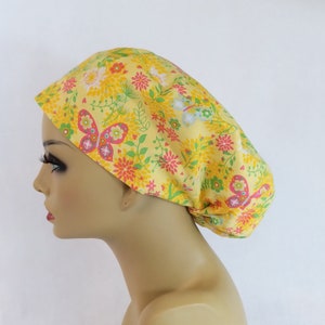 May include: A yellow floral patterned scrub cap featuring butterflies, flowers, and foliage in pink, green, and white. The cap is made of a soft fabric and is designed to cover the head.