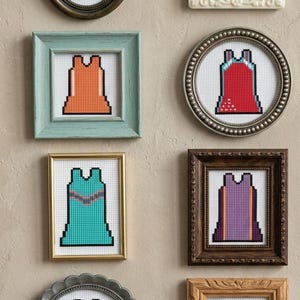 Op de afbeelding: Verzameling ingelijste kruissteekkunstwerken, elk met een gepixeld jurkontwerp. De jurken zijn in verschillende kleuren, waaronder blauw, roze, oranje, rood, blauwgroen, paars, geel en groen, weergegeven in verschillende framestijlen.