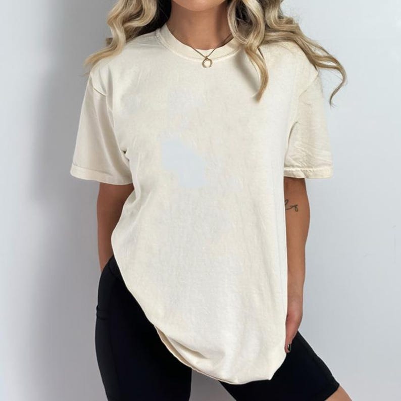 May include: Cream-colored, short-sleeved t-shirt. The shirt has a slightly oversized fit and a crew neck. The fabric appears soft and slightly textured. The model is wearing black shorts. The overall style is casual and relaxed.