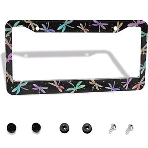 May include: A rectangular black license plate frame featuring a repeating pattern of watercolor-style dragonflies in shades of teal, purple, and peach, accented with small yellow dots. The set includes two black screw covers, two washers, and two screws.