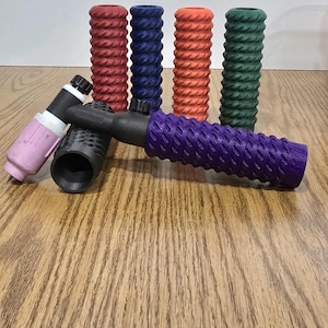 Op de afbeelding: Een verzameling laspistoolhandvatten in verschillende kleuren, waaronder rood, blauw, oranje, groen, zwart en paars. De handvatten hebben een gestructureerd, geribbeld ontwerp voor grip en zijn gerangschikt op een houten oppervlak.