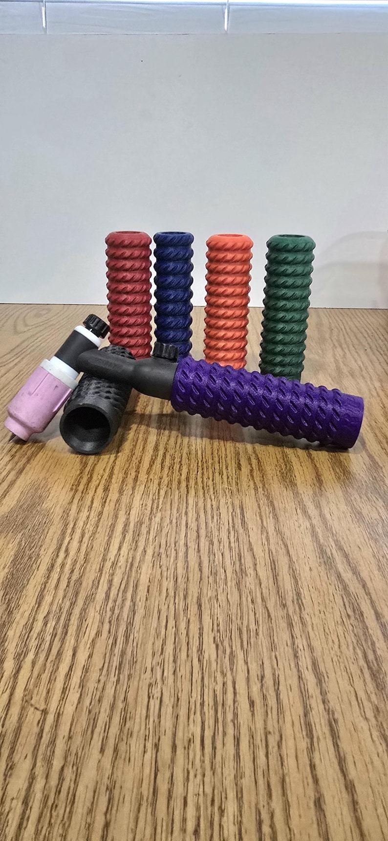 Op de afbeelding: Een verzameling laspistoolhandvatten in verschillende kleuren, waaronder rood, blauw, oranje, groen, zwart en paars. De handvatten hebben een gestructureerd, geribbeld ontwerp voor grip en zijn gerangschikt op een houten oppervlak.
