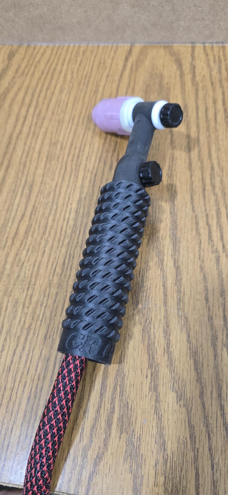 Op de afbeelding: Een zwarte TIG-lastoorts met een getextureerde handgreep en een rood-zwart patroon kabel. De toortskop is zwart en wit met een roze keramische nozzle.