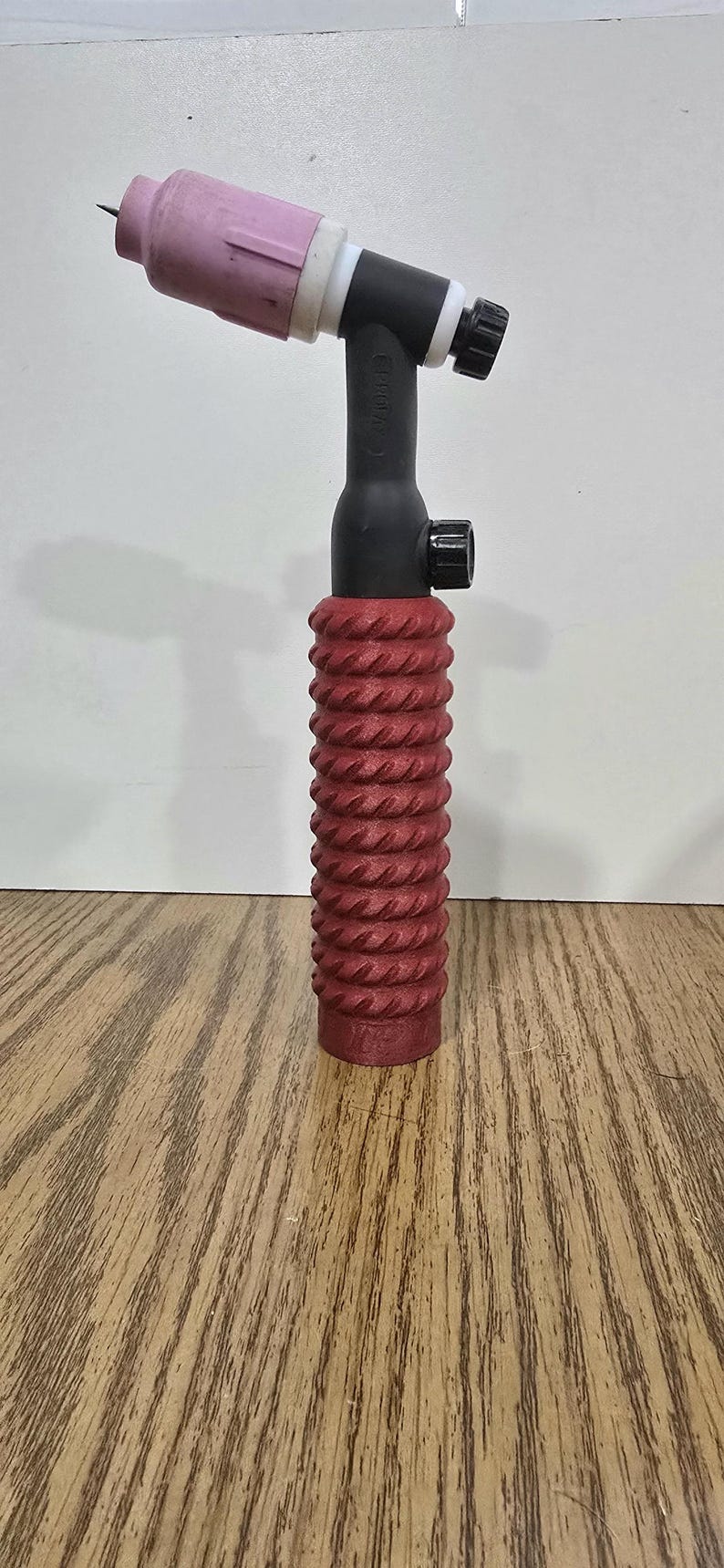 Op de afbeelding: Een TIG-lastoorts met een rode, getextureerde handgreep, zwarte behuizing en een roze keramische nozzle. De toorts heeft een zwarte knop en een punt. De achtergrond is licht.