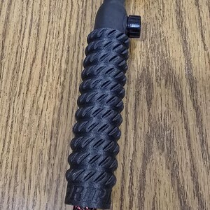 May include: A black TIG welding torch with a textured handle. The torch has a pink ceramic nozzle, a black control knob, and a red and black patterned cable. The handle has a ridged design for grip.