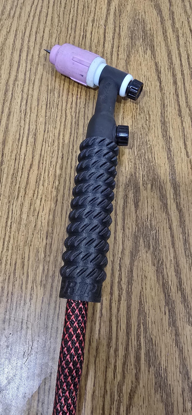 May include: A black TIG welding torch with a textured handle. The torch has a pink ceramic nozzle, a black control knob, and a red and black patterned cable. The handle has a ridged design for grip.