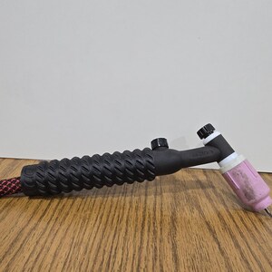 May include: A black TIG welding torch with a textured handle and a pink ceramic nozzle. The torch is connected to a red and black cable. The torch is on a wooden surface.