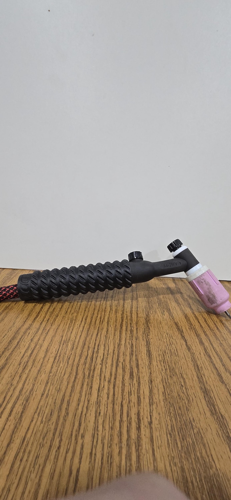 May include: A black TIG welding torch with a textured handle and a pink ceramic nozzle. The torch is connected to a red and black cable. The torch is on a wooden surface.