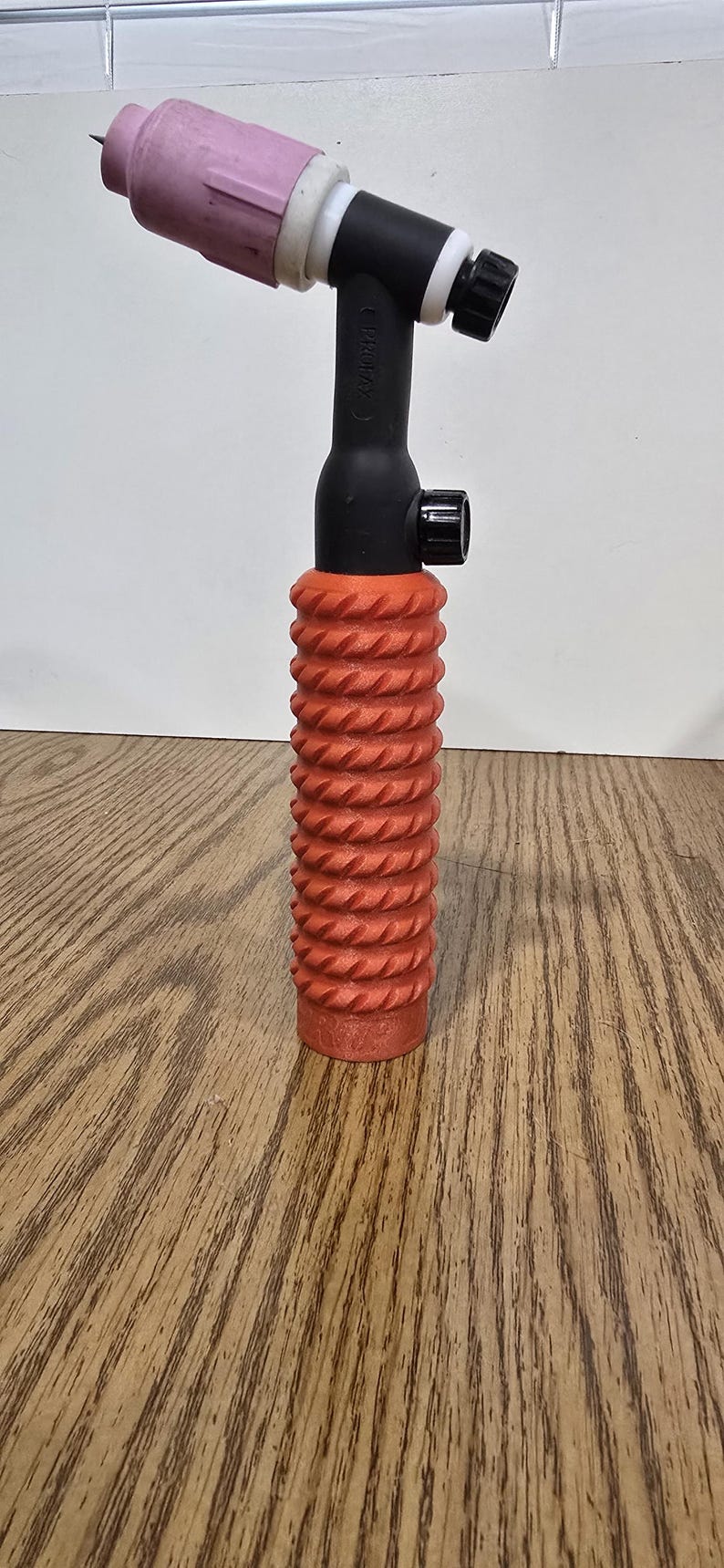 Op de afbeelding: Een TIG-lastoorts met een roze keramische nozzle, zwarte behuizing en een oranje, getextureerde handgreep. De toorts is ontworpen voor precisielassen en staat tegen een witte achtergrond en houten oppervlak.