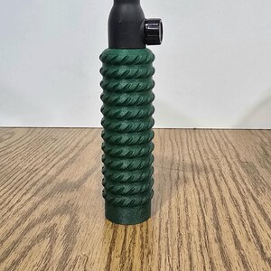 Op de afbeelding: Een lastoorts met een groene, getextureerde handgreep, een zwarte behuizing en een roze keramische nozzle. De toorts staat tegen een lichte achtergrond en een houten oppervlak.