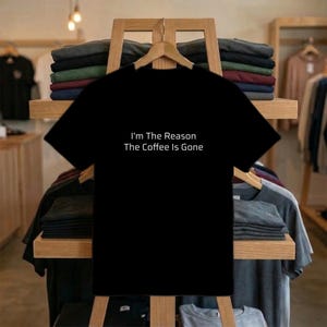 May include: Black t-shirt with the white text "I'm The Reason The Coffee Is Gone". The tee is displayed on a wooden easel in a shop, with folded shirts on shelves behind it. The shop has a wooden interior.
