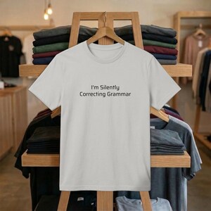 May include: A light gray t-shirt with the text "I'm Silently Correcting Grammar" in black. The shirt is hanging on a wooden rack in a clothing store. Other folded shirts are visible on the shelves.