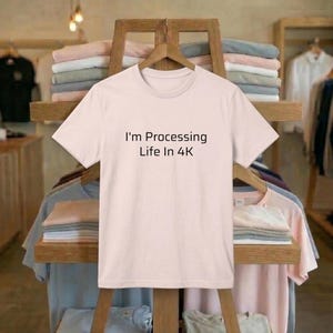 May include: A pale pink t-shirt displaying the text "I'm Processing Life In 4K" in black. The shirt is suspended on a wooden clothes rack, with folded garments visible in the background. The t-shirt has a round neckline.