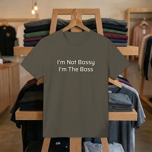 May include: An olive green t-shirt with the white text "I'm Not Bossy I'm The Boss". The t-shirt is displayed on a wooden rack in a shop, with other folded and hanging clothes visible in the background.