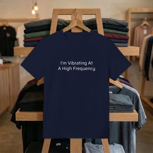 May include: Navy blue t-shirt with the white text "I'm Vibrating At A High Frequency." The shirt is displayed on a wooden rack with other folded shirts. The background shows a clothing store setting.
